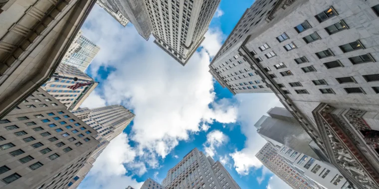 A worm's-eye view of tall, grey stone buildings in a city, with a bright blue sky and white clouds visible between them.